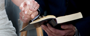 Nærbillede af et par med vielsesringe, der holder hinanden i hånden over en åben bibel.