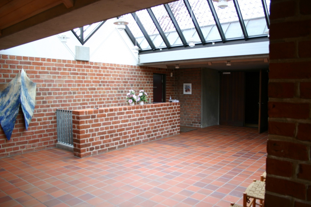 Modernes Kirchenfoyer mit roten Backsteinwänden, Fliesenboden, Dachfenster und Wandskulptur.